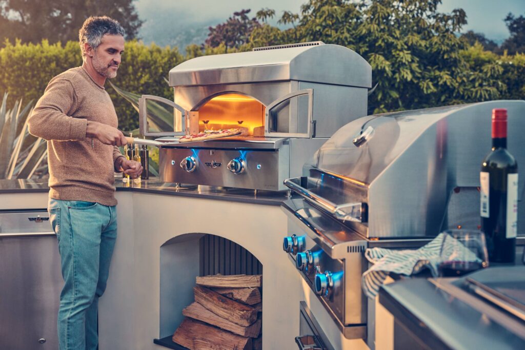 Man cooking pizza in an outdoor wood-fired pizza oven built into a backyard kitchen island