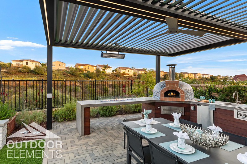 Outdoor kitchen with brick pizza oven under louvered pergola overlooking hillside neighborhood