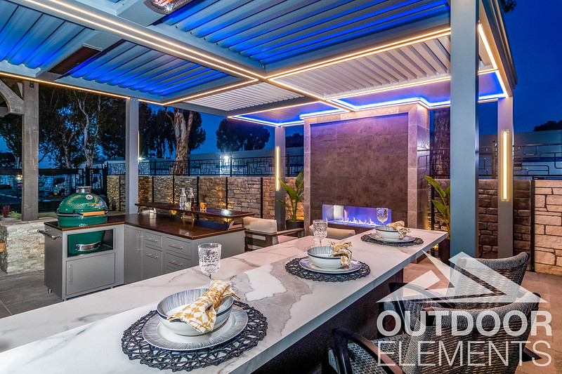 Pergola with blue and amber LED lighting over dining table and stone fireplace at night