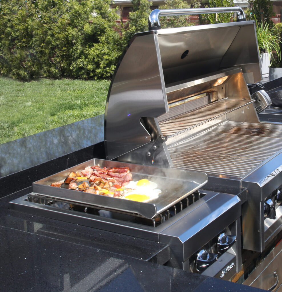 Bacon, eggs, and vegetables cooking on stainless steel griddle attachment on outdoor grill