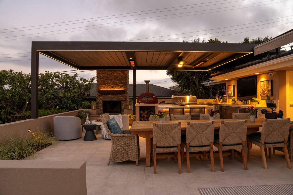Outdoor kitchen pavilion at dusk with pizza oven, dining table and string lighting