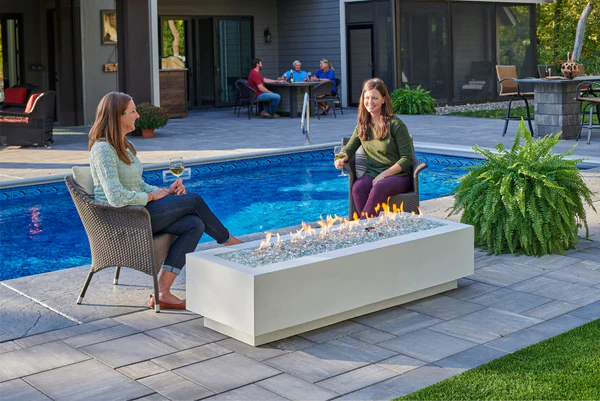 Two women relaxing by a poolside gas fire table on a backyard patio at dusk