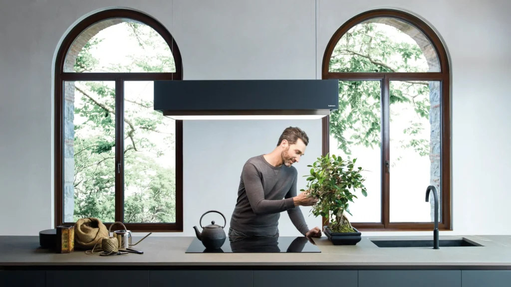 Man tending a bonsai tree on a kitchen island beneath arched black-framed windows