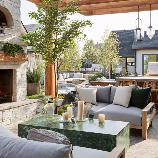 Outdoor living space with stone fireplace, pizza oven, wood pergola, gray sofa seating, green marble coffee table, and built-in kitchen with bar stools