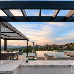 Pergola-covered outdoor kitchen bar with wine glasses overlooking infinity pool at sunset