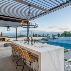 Outdoor kitchen island with waterfall countertop and rattan bar stools overlooking infinity pool at dusk