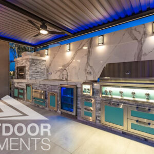 Outdoor kitchen with marble backsplash, teal cabinetry, and pizza oven under blue-lit pergola at night