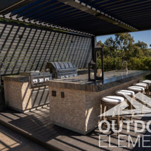 Stone outdoor kitchen with grill and bar seating under slatted pergola trellis