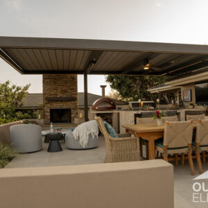 Pergola-covered patio with stone fireplace, pizza oven, and outdoor dining table at sunset