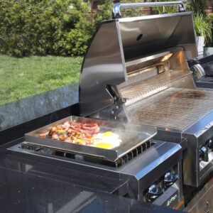 Bacon, eggs, and vegetables cooking on stainless steel griddle attachment on outdoor grill